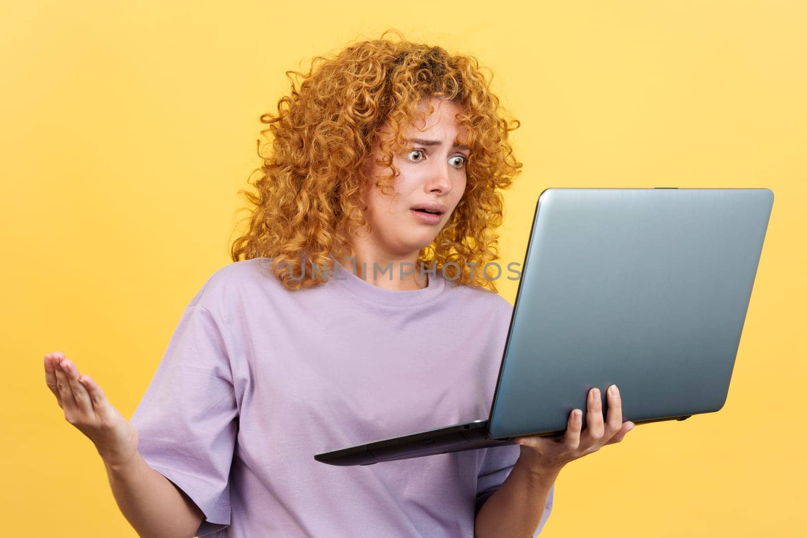Studio image with yellow background of a worried woman with curly hair standing while holding a laptop expressing a problem