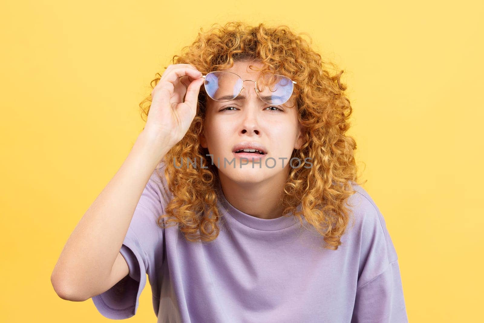 Studio image with yellow background of a young woman taking off her glasses to see far away while looking at camera
