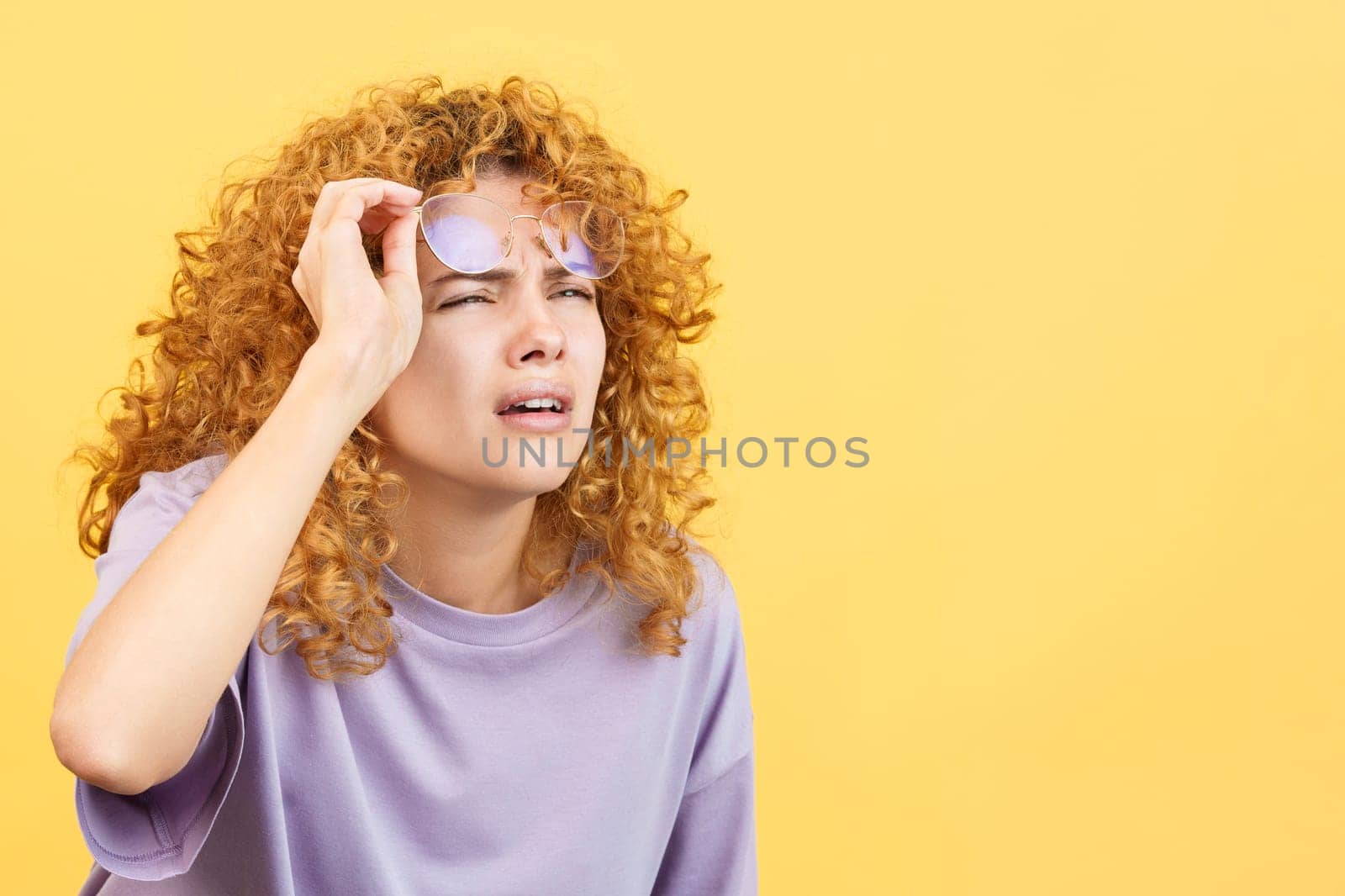 Studio image with yellow background of a woman with curly hair taking off her glasses to see far away but finds it hard to do so