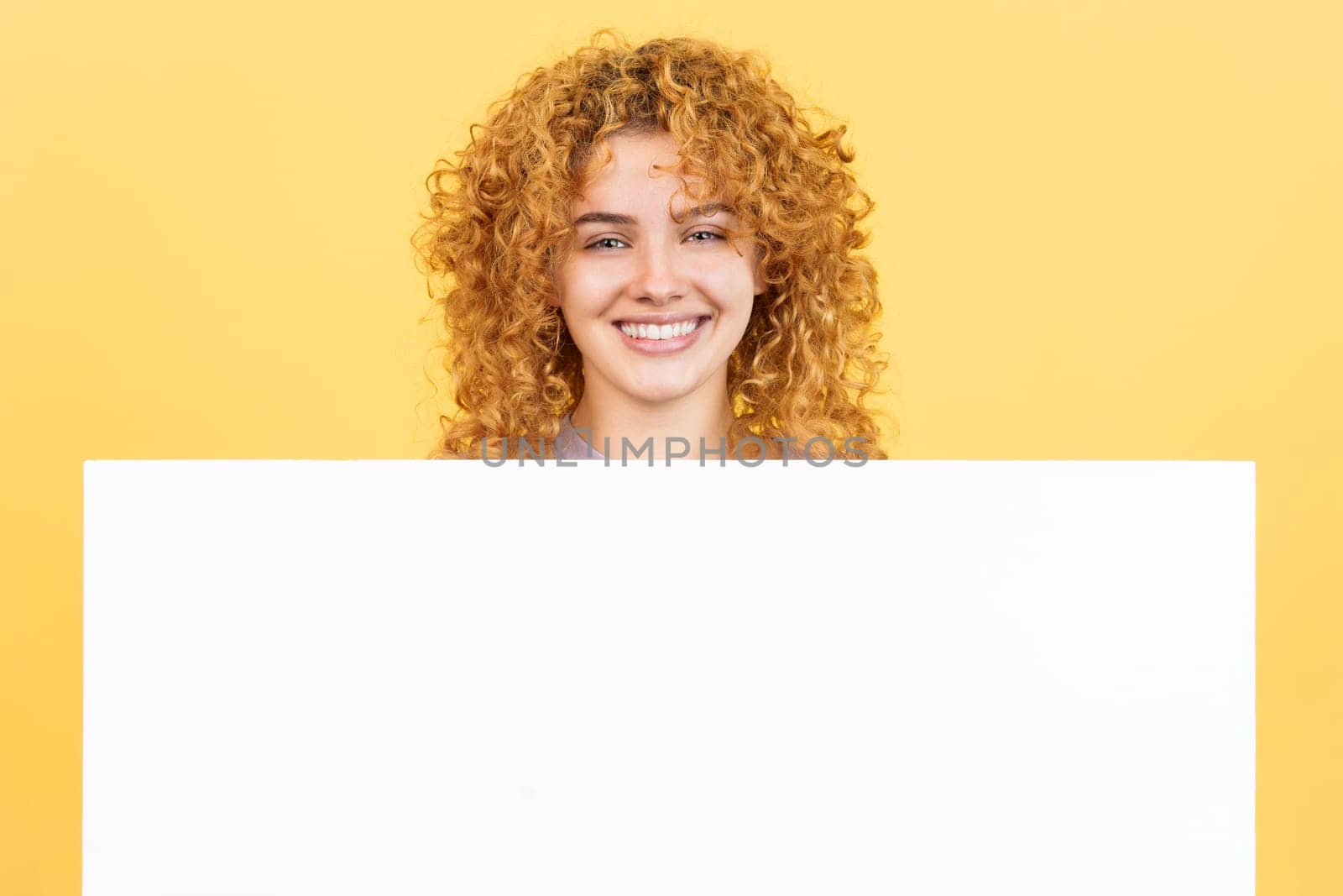 Studio image with yellow background of a beauty and smiley woman with curly hair holding a blank panel