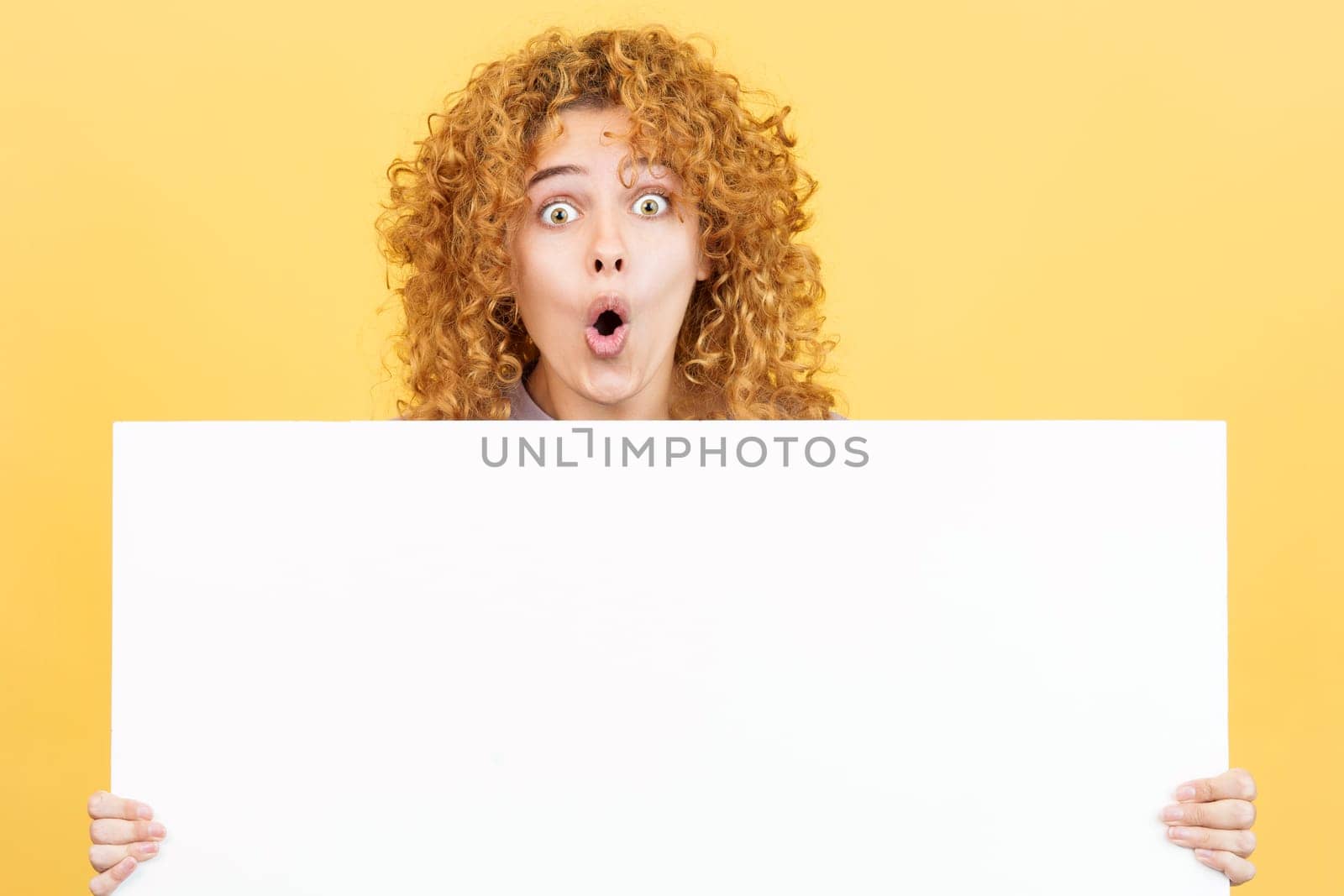 Studio image with yellow background of a woman with curly hair holding a blank panel while expressing surprise