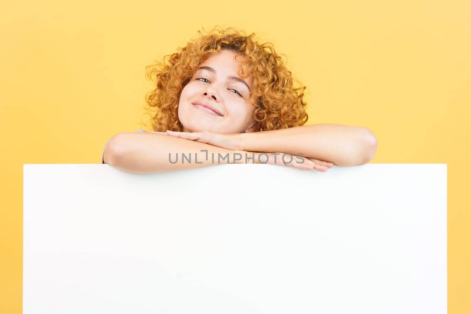 Studio image with yellow background of a relaxed woman with curly hair leaning on a blank panel