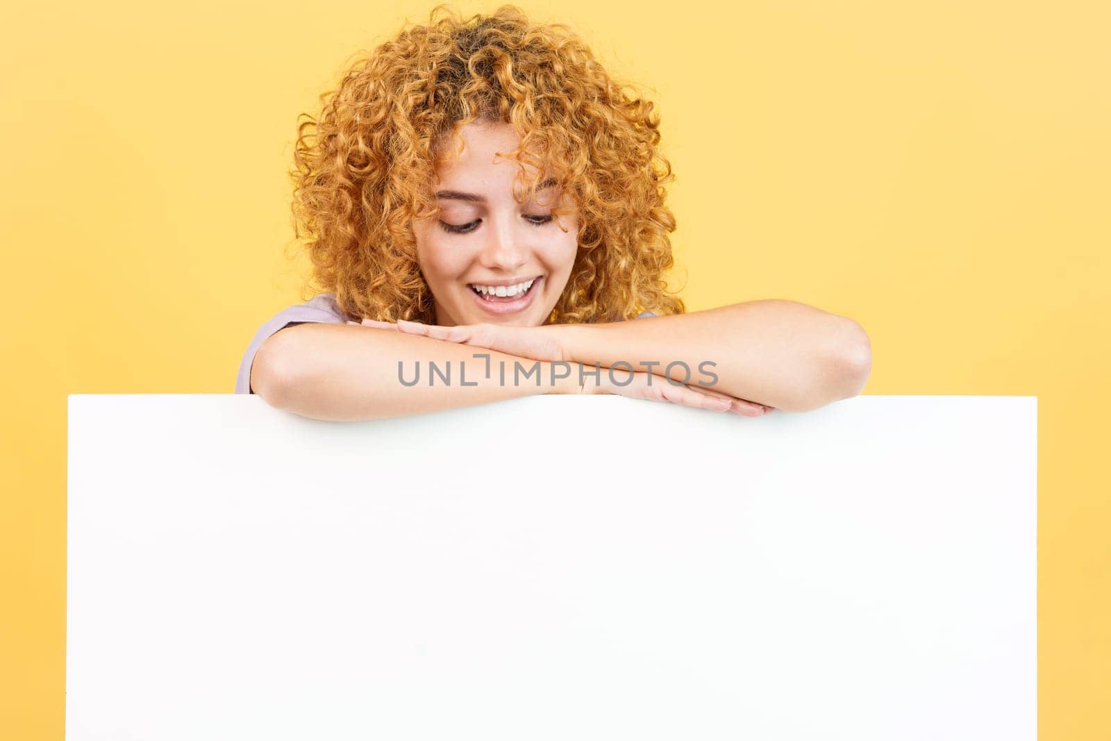 Studio image with yellow background of a relaxed woman with curly hair leaning and looking down to a blank panel