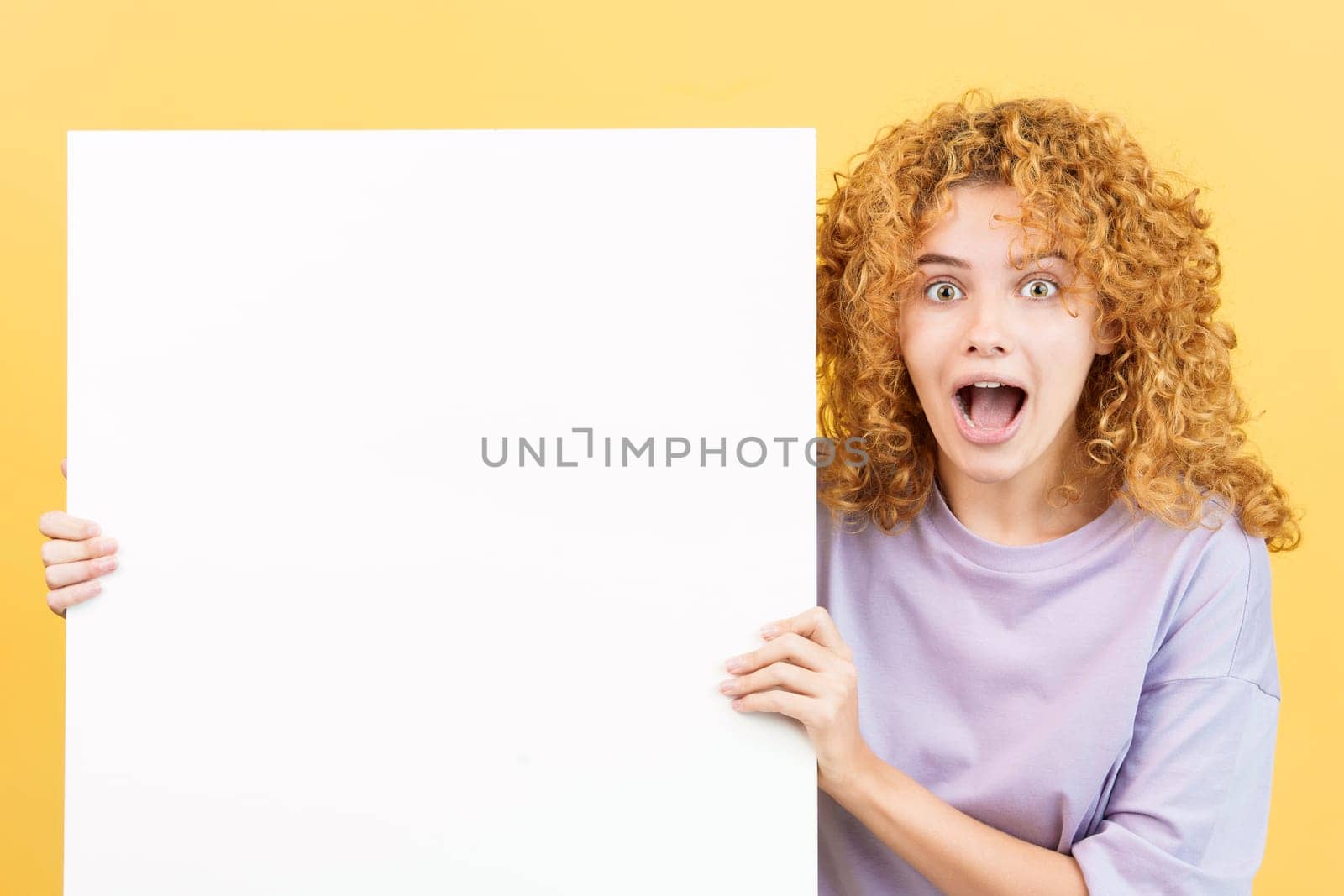 Studio image with yellow background of a beauty surprised woman with curly hair holding a blank board
