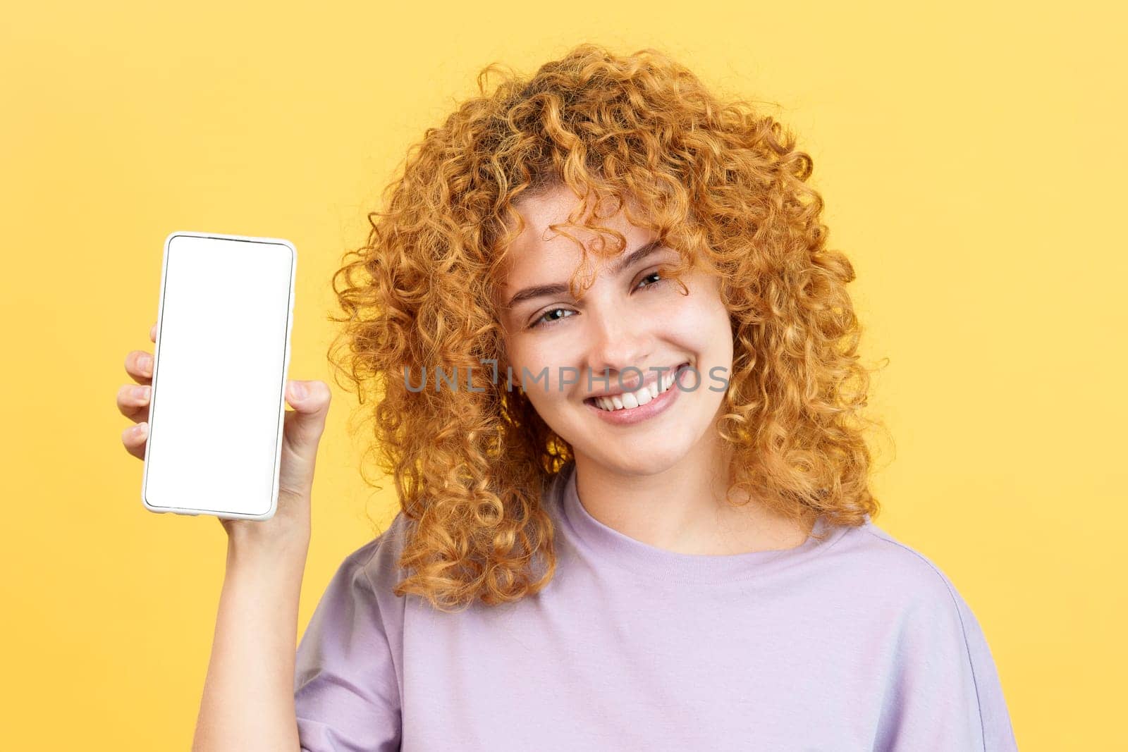 Studio image with yellow background of a smiley woman looking at camera while holding a mobile with the blank screen