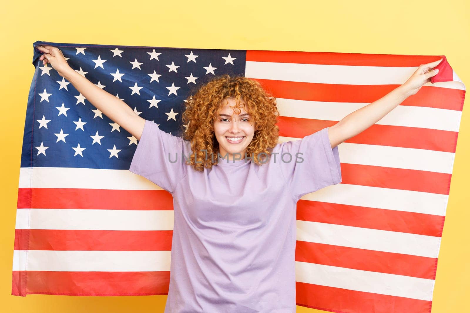 Studio image with yellow background of a beauty and happy woman with curly hair raising a north america national flag