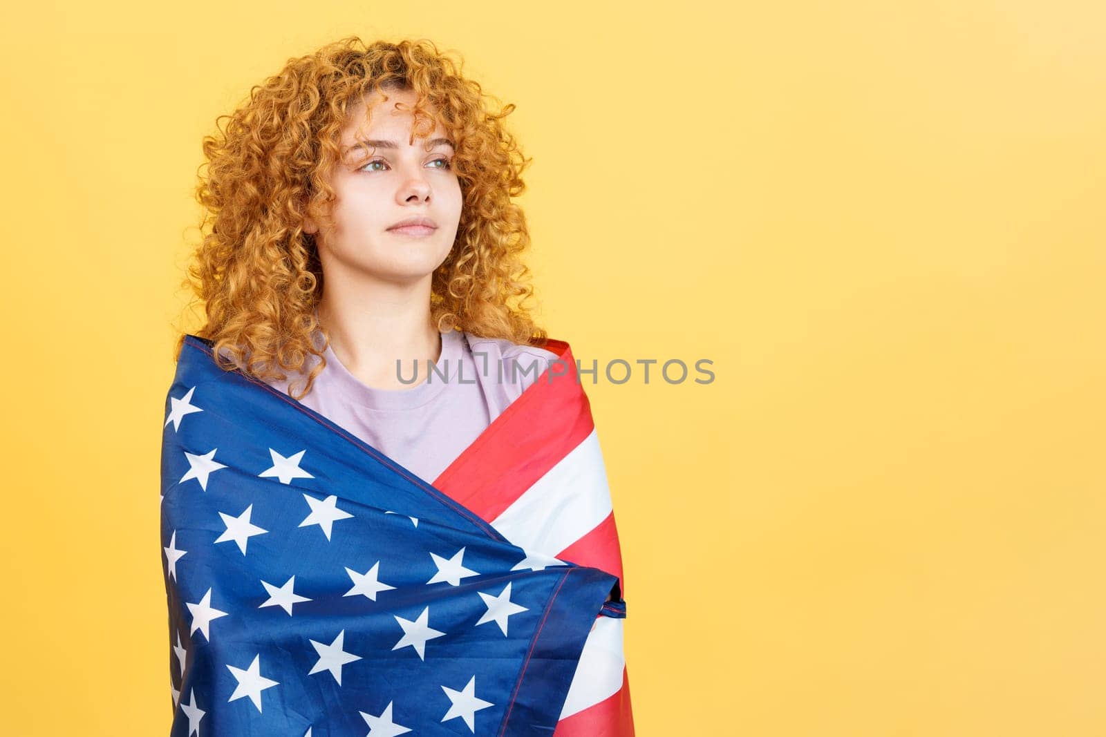 Studio image with yellow background of a beauty and distracted serious woman with curly hair wrapping with a north america national flag