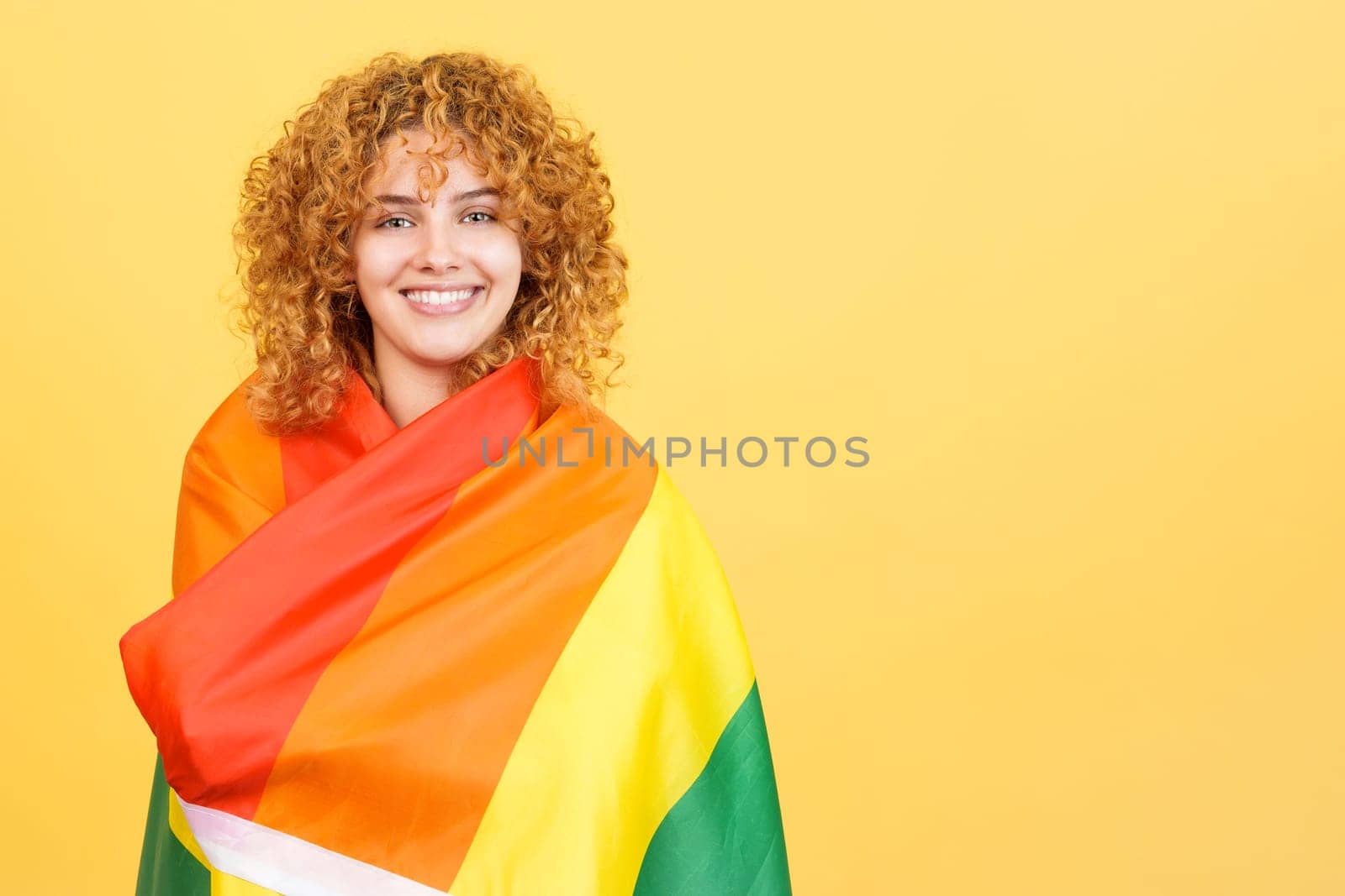 Studio image with yellow background of a woman wrapped with a lgbt flag looking at camera