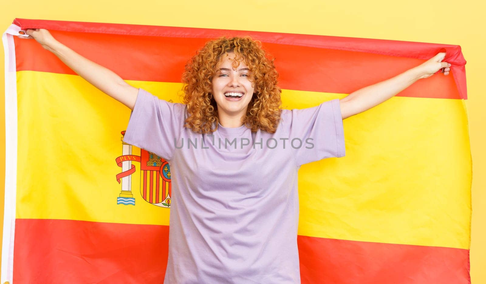 Studio image with yellow background of a happy beauty woman with curly hair raising a spanish national flag