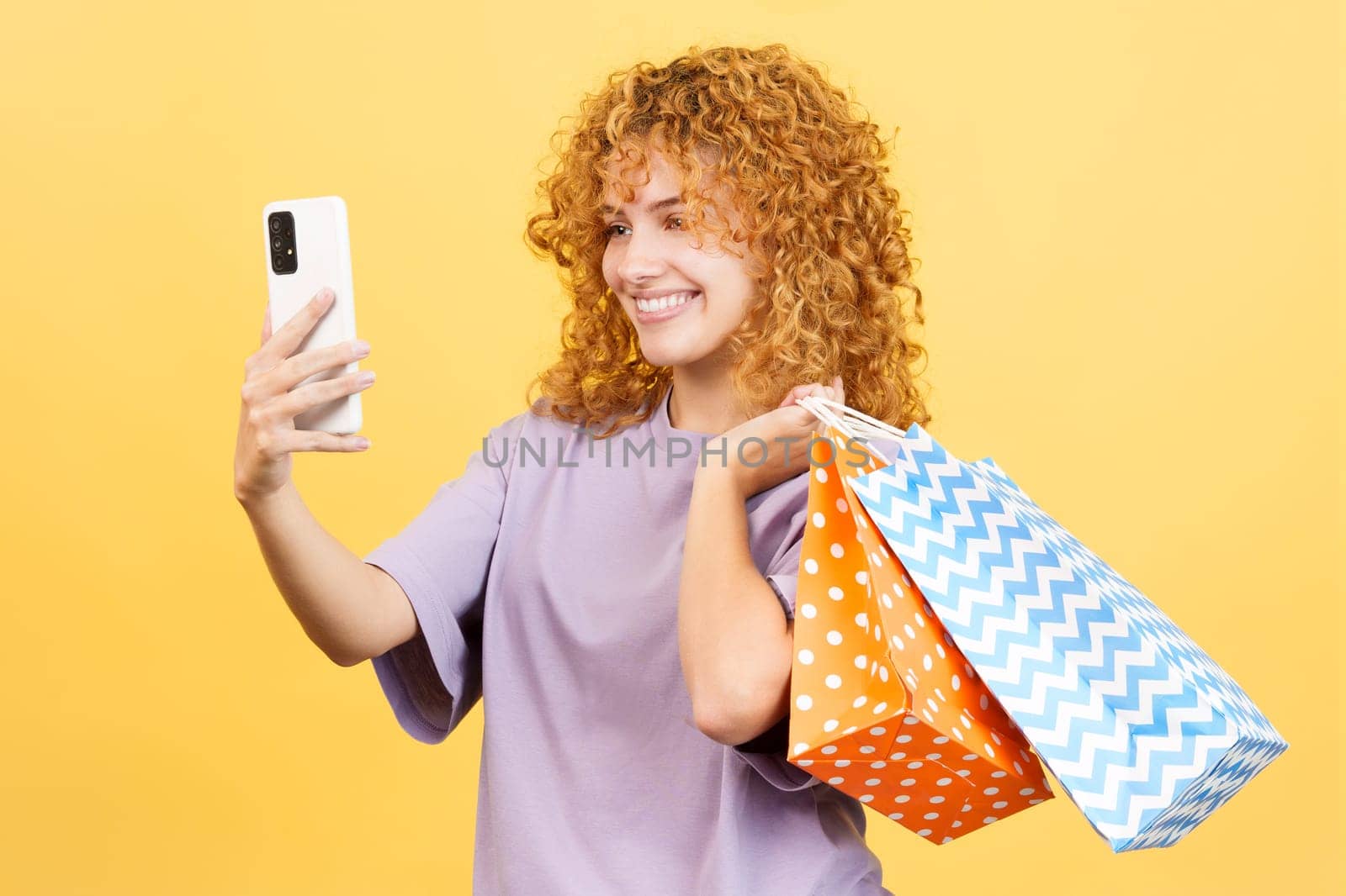Studio image with yellow background of a happy young woman with curly hair taking a selfie while holing shopping bags