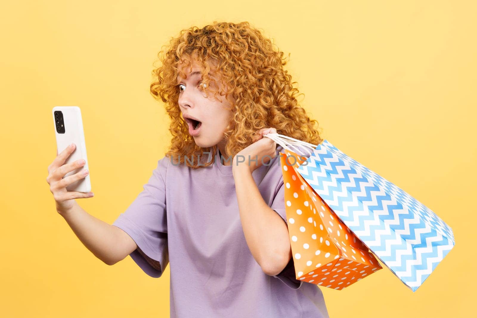 Studio image with yellow background of a young woman with facial expression of surprise using the mobile while holing shopping bags