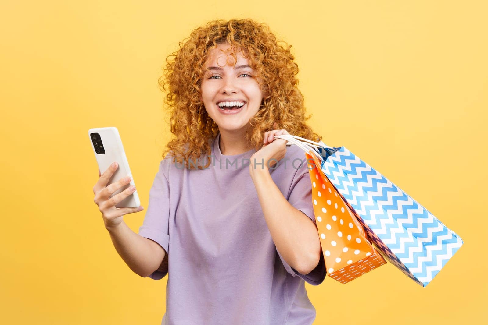 Studio image with yellow background of a young smiley woman with curly hair using the mobile while holing shopping bags