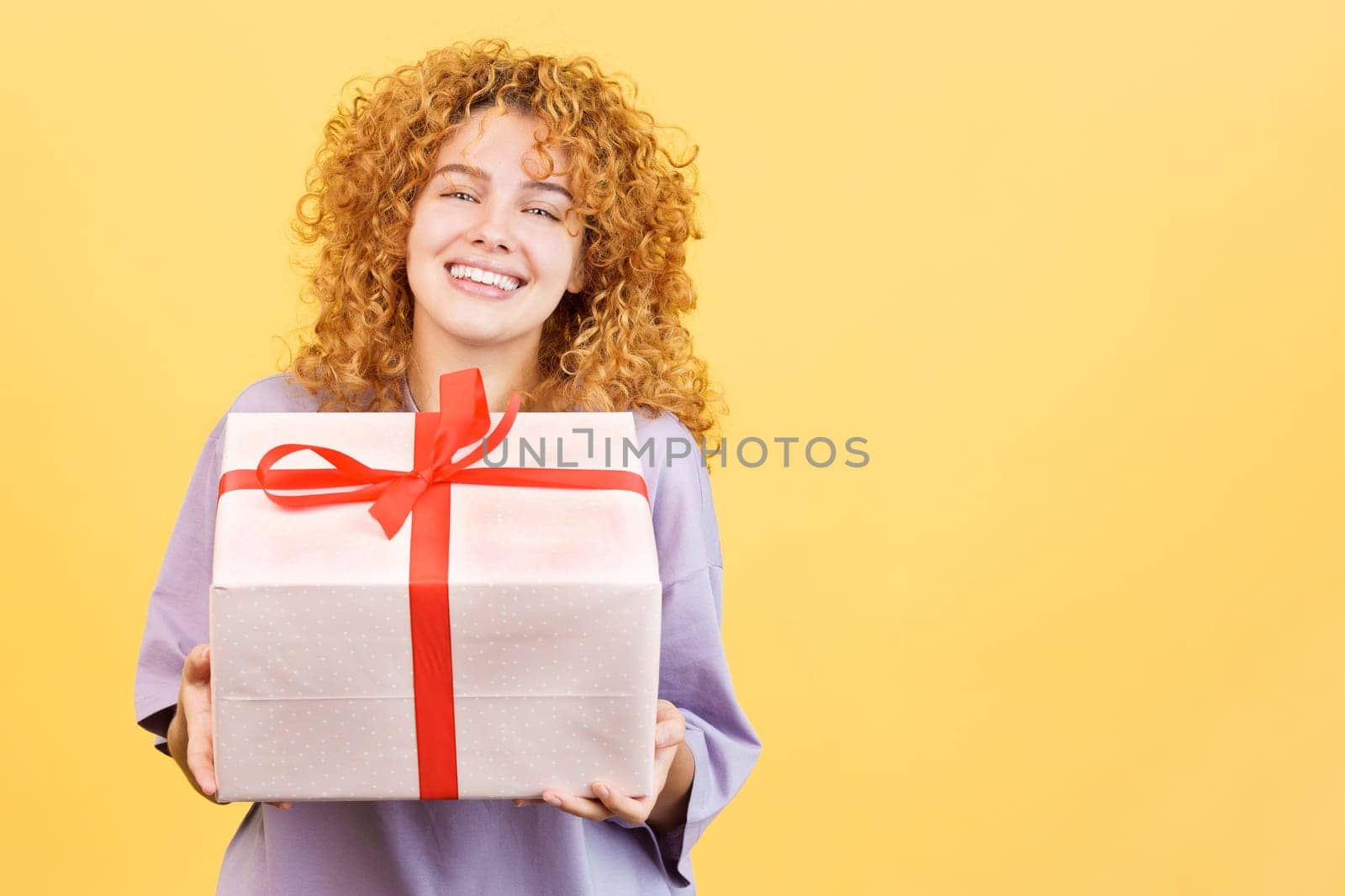 Studio image with yellow background of a happy young woman smiling at camera while giving a gift