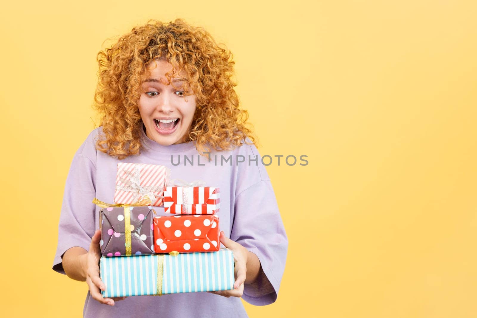 Studio image with yellow background of an excited young woman with curly hair and casual clothes receiving many gifts