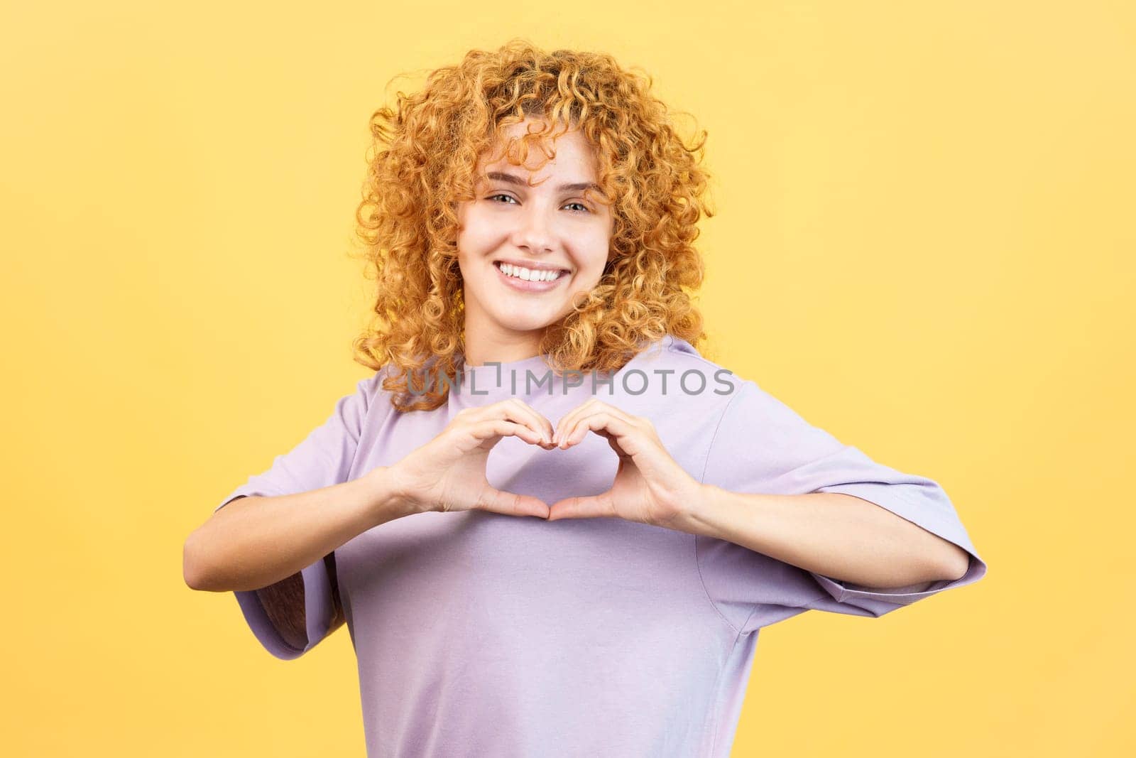 Studio image with yellow background of a happy young woman with curly hair representing a heart in the shape of fingers