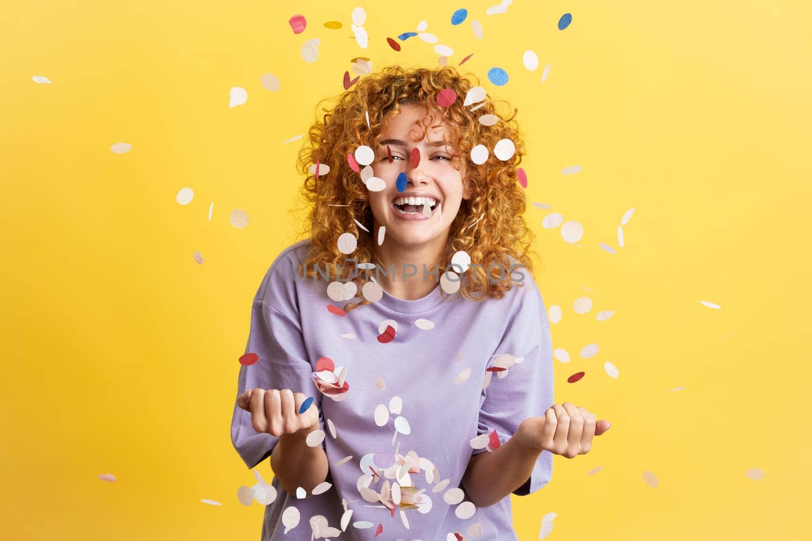 Studio image with yellow background of a young and cheerful woman with curly hair surrounded by confetti in the air