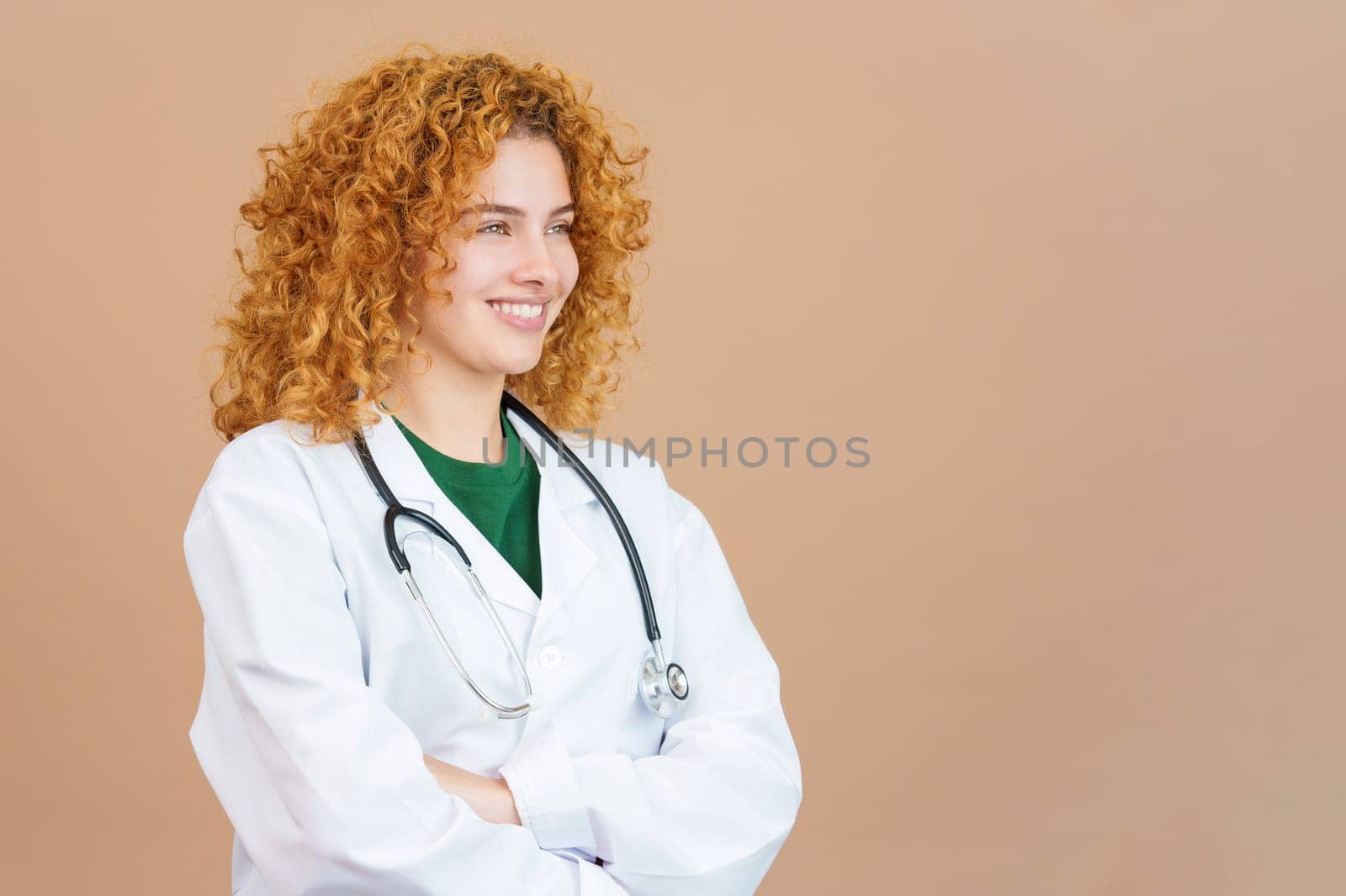 Studio image of a female doctor with curly hair smiling while looking aside with the arms crossed