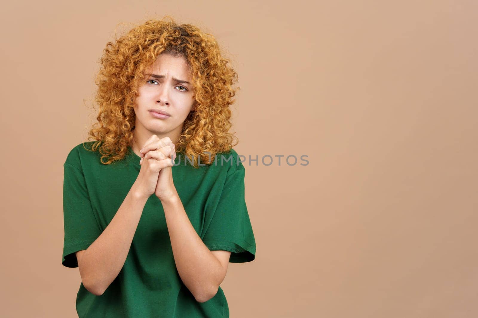 Studio photo of woman beggong with curly hair and a worried face, praying with her hands clasped while looking into the camera