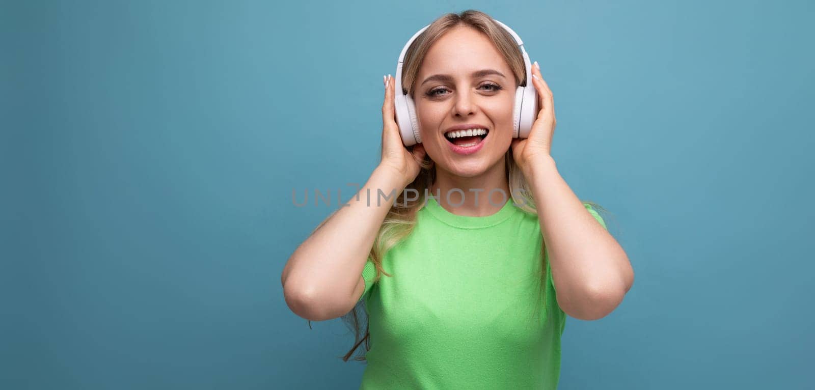 close-up of an adorable blond girl in a casual outfit relaxing and dancing in big white headphones on a blue background by TRMK