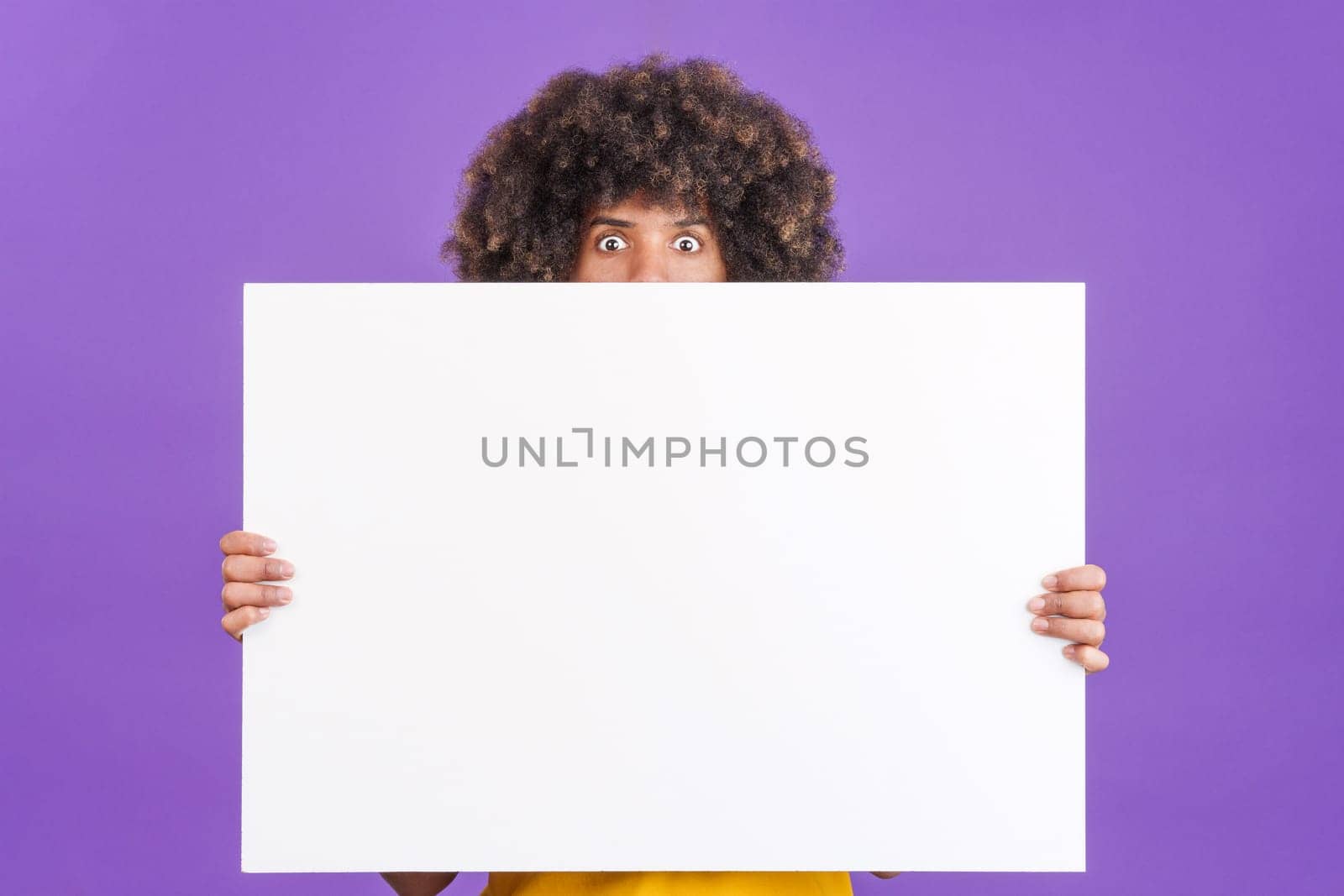 Surprised african man with curly hair hiding behind a blank panel in studio