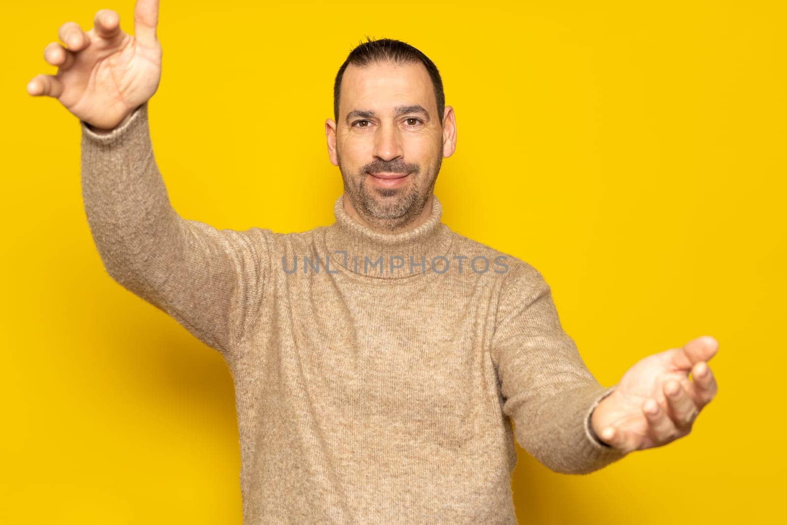 Bearded Hispanic man wearing a beige turtleneck in a friendly attitude looking at the camera smiling with open arms for a hug. Cheerful expression embracing happiness. Isolated on yellow background