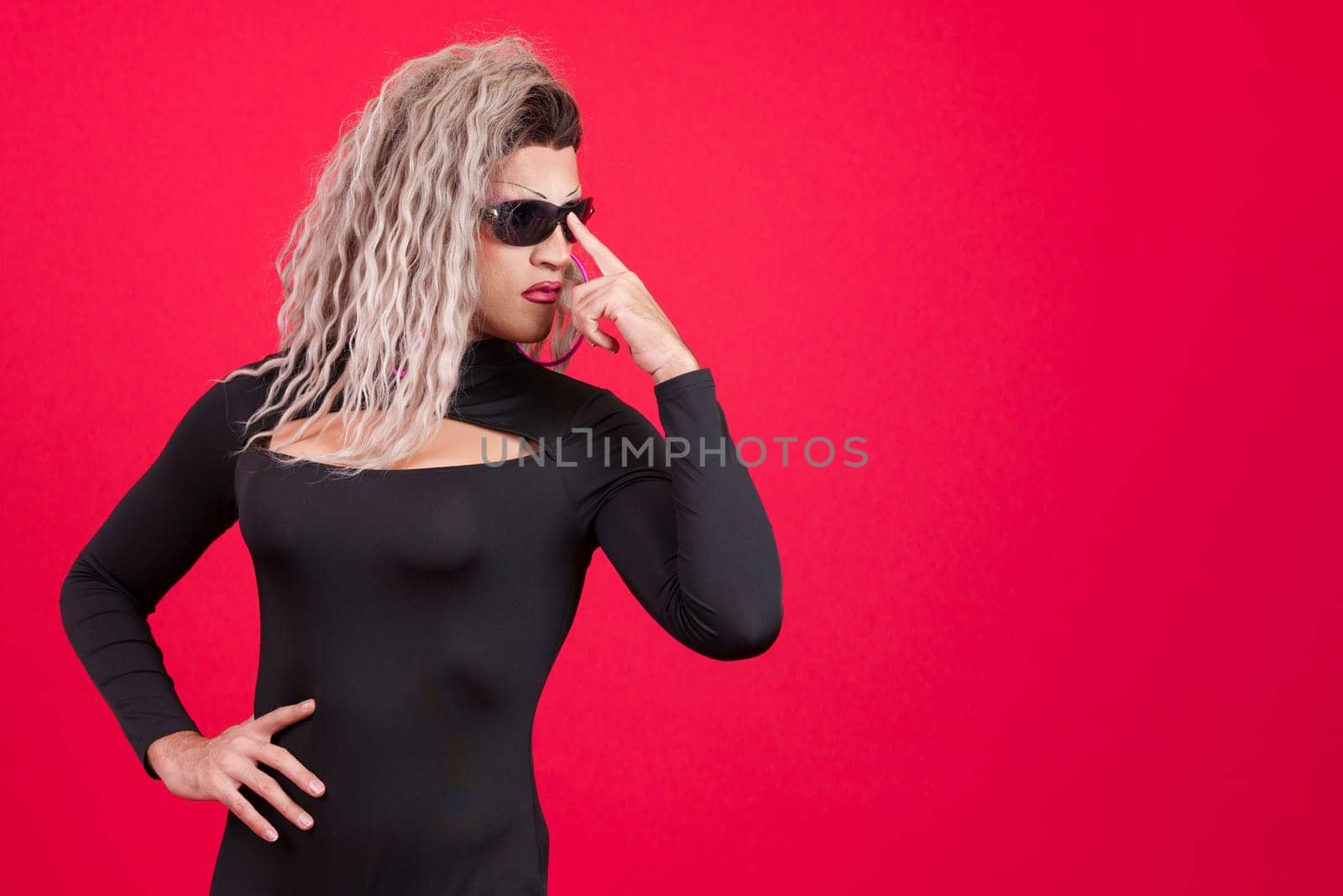 Transgender person dressed as drag queen wearing sunglasses posing with rebel attitude in studio with red background