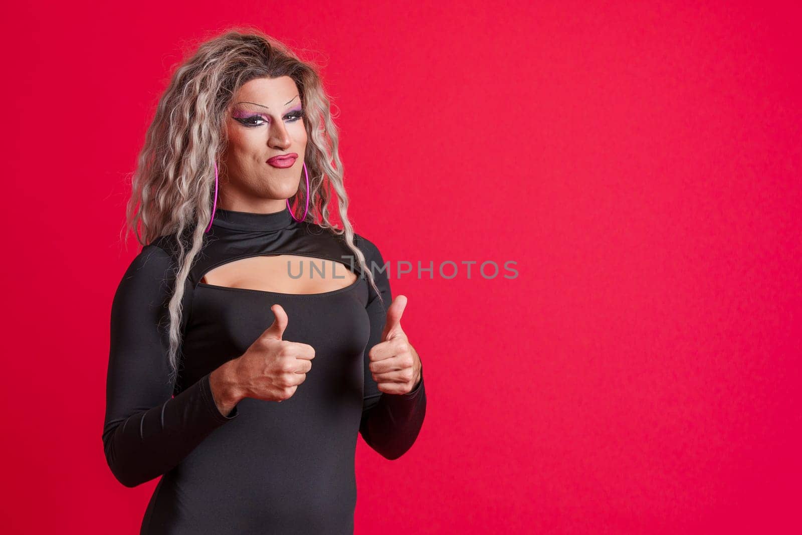 Transgender person with make up gesturing agreement raising thumbs up in studio with red background