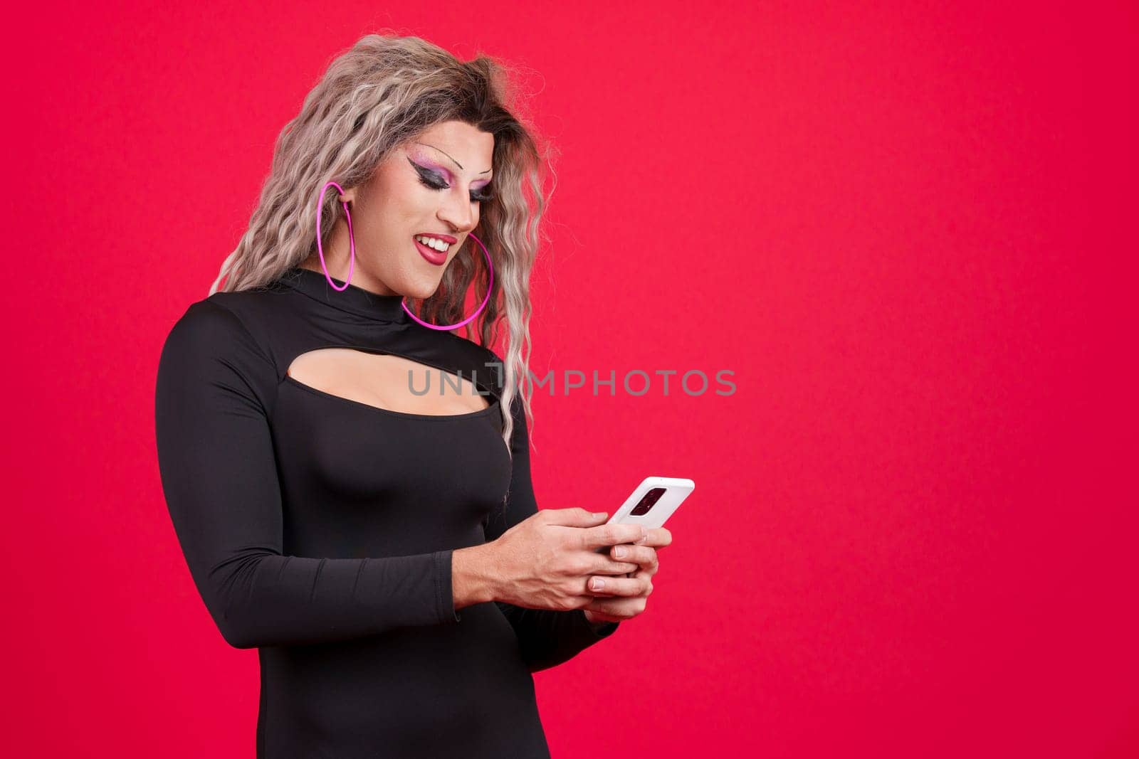 Happy transgender person dressed as drag queen smiling while using a mobile in studio with red background