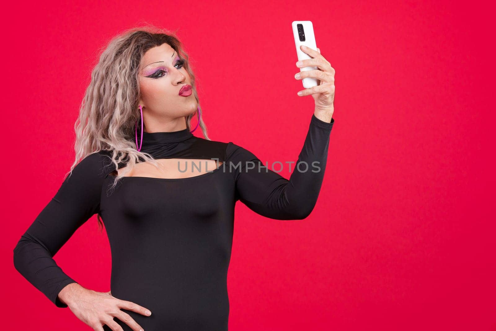 Sensual transgender person with make up kissing while taking a selfie with a mobile phone in studio with red background