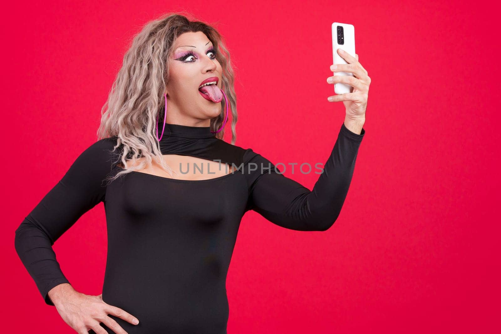 Playful transgender person taking a selfie with a mobile phone while grimacing in studio with red background