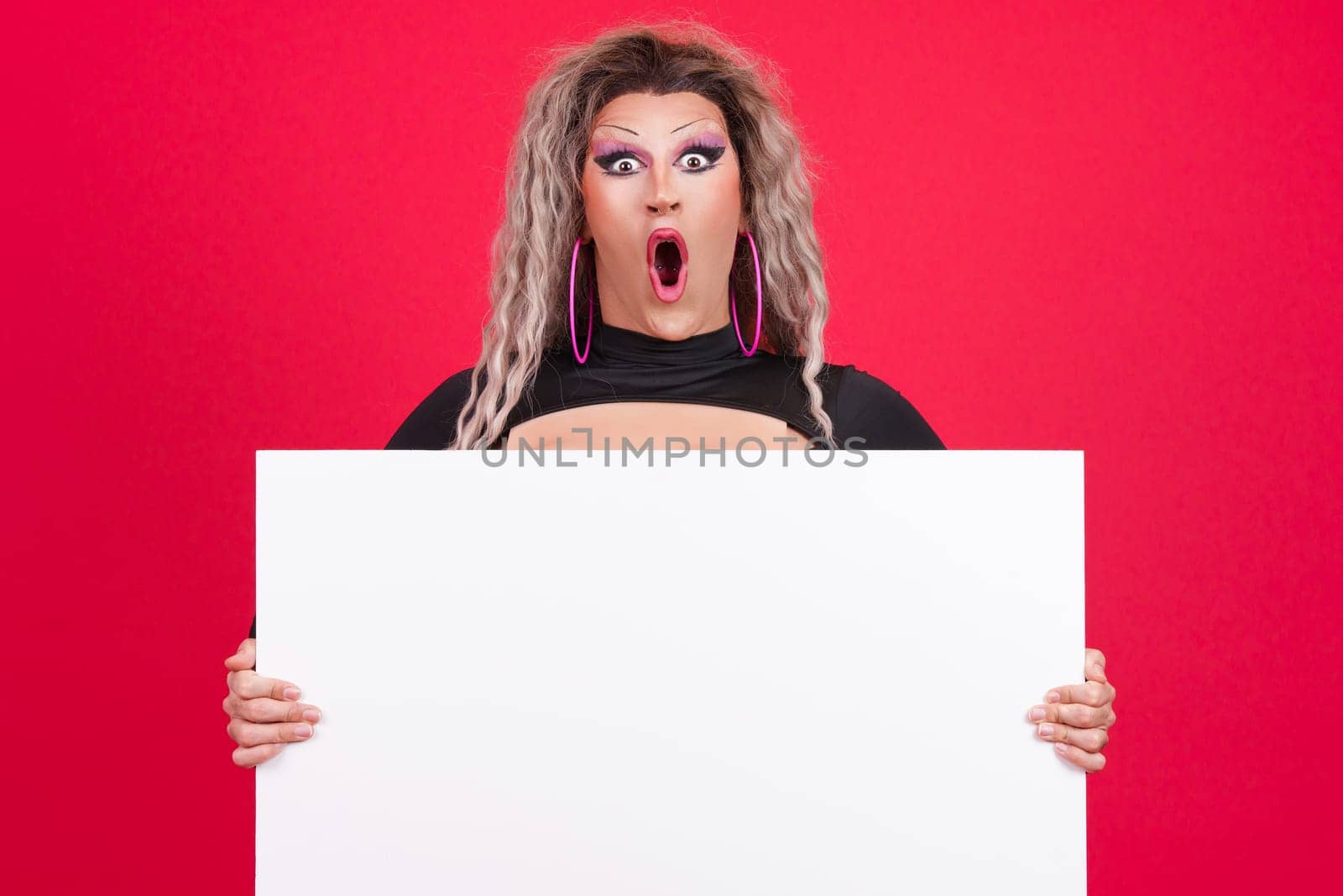 Surprised transgender person with make up opening the mouth while holding a blank panel in studio with red background