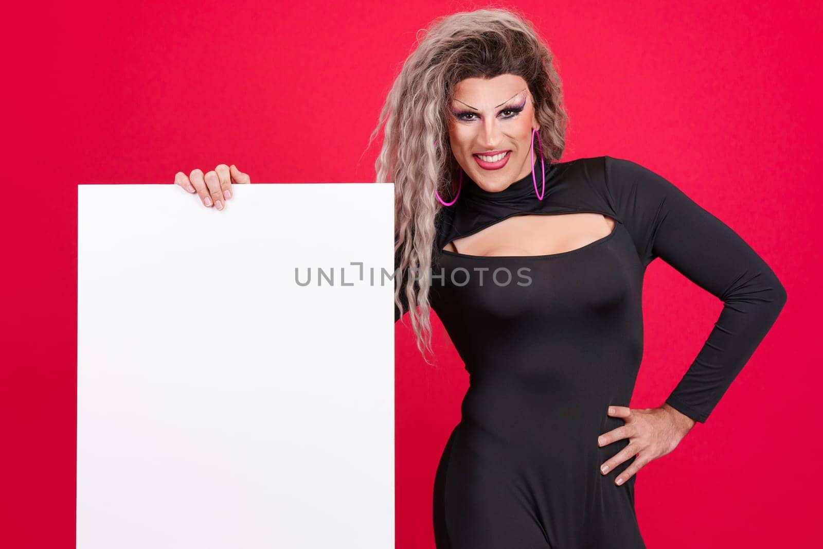 Smiley transgender person dressed as drag queen and with make up holding a blank panel in studio with a red background