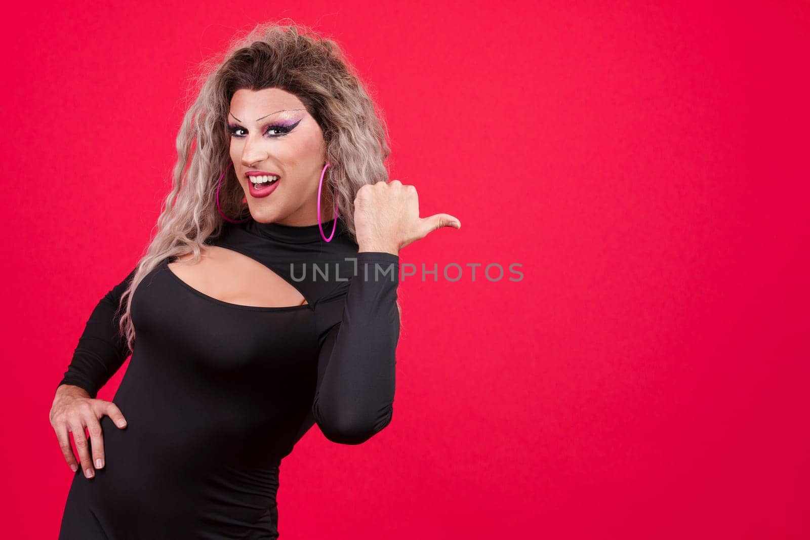 Transgender person dressed as drag queen smiling at camera and pointing to the side in studio with red background