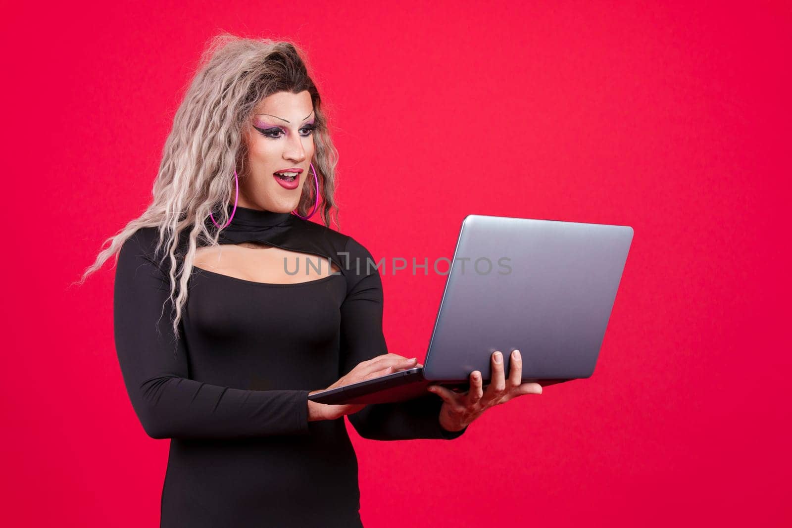 Surprised transgender person dressed as drag queen standing and using a laptop in studio in red background