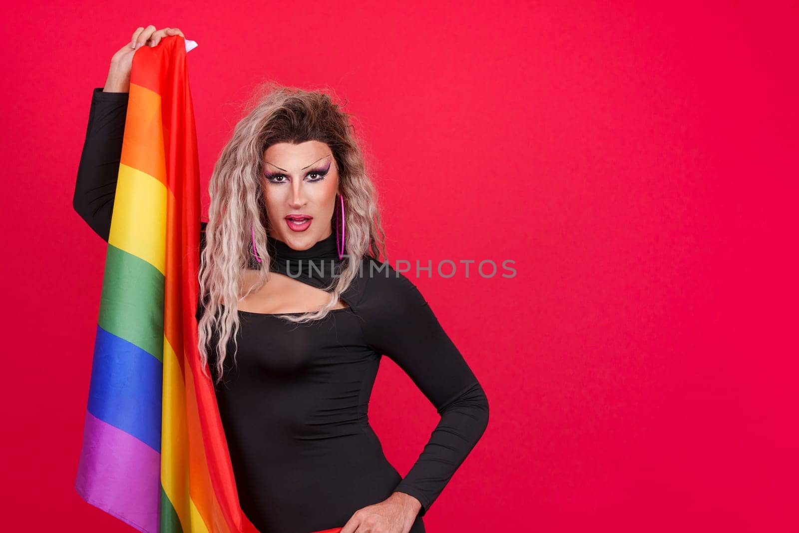 Transgender person dressed as drag queen looking at camera with a rainbow lgbt flag in studio with red background
