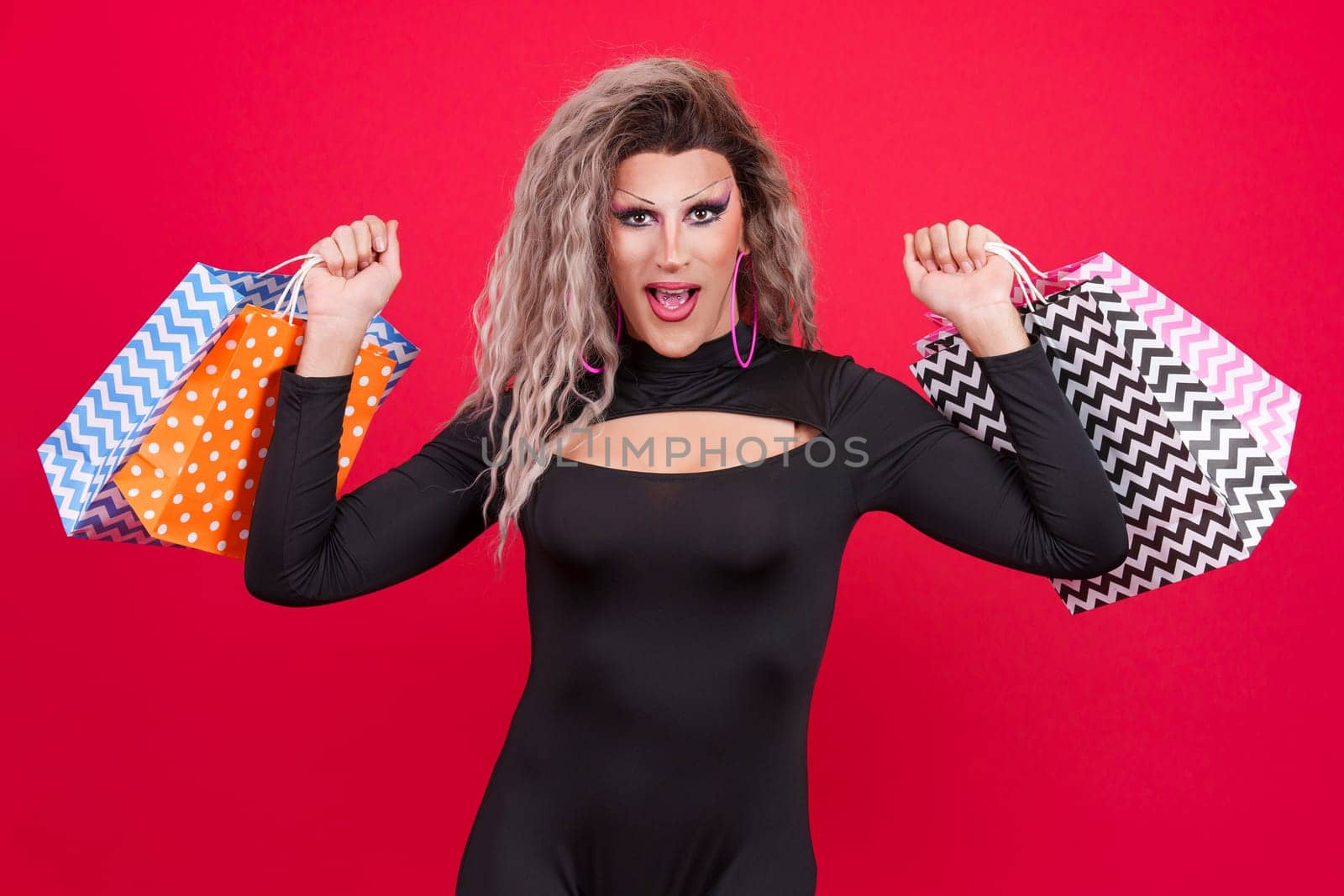 Sensual transgender person dressed as drag queen smiling at the camera while showing shopping bags in studio with red background