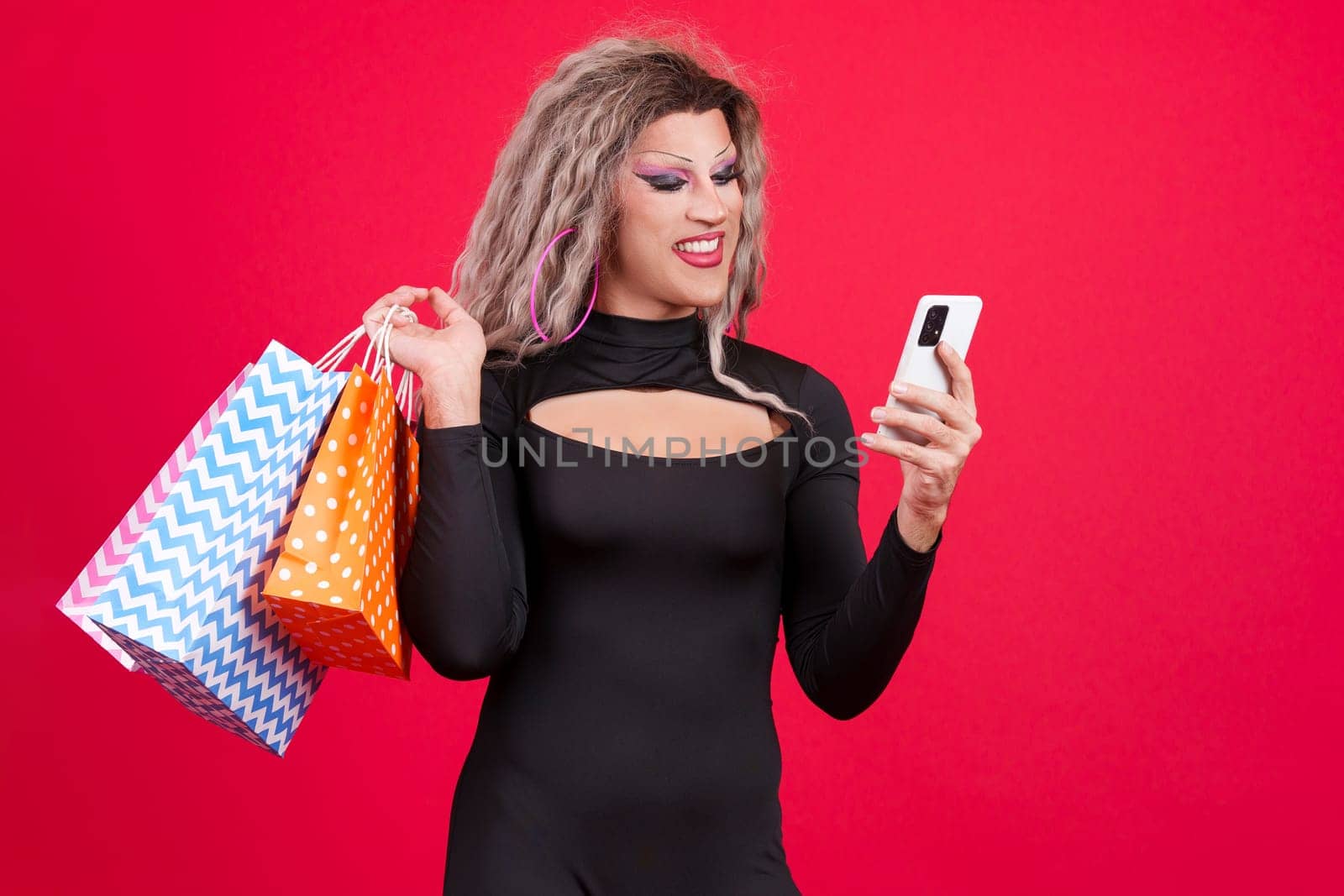 transgender person dressed as drag queen using a mobile while holding shopping bags in studio with red background