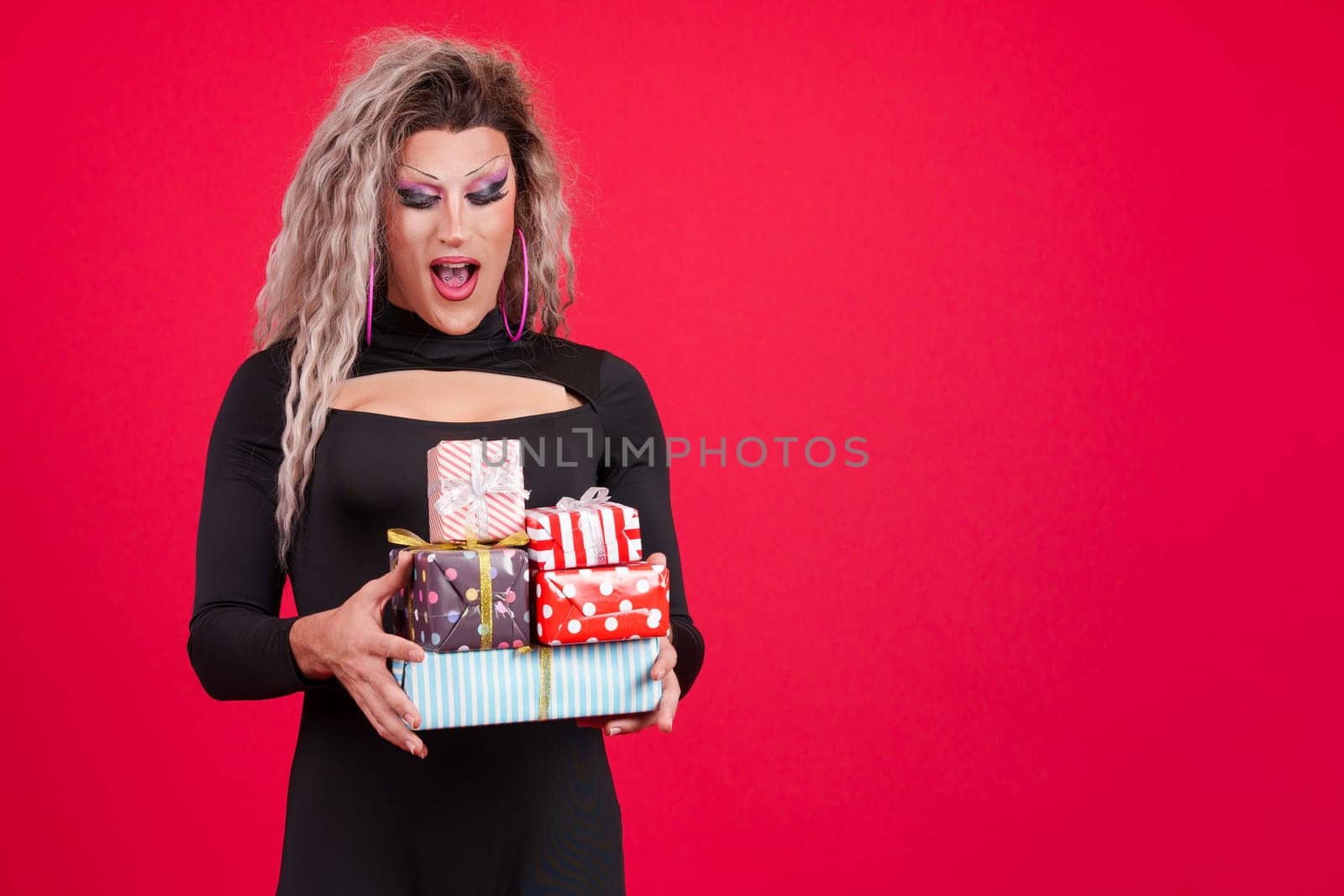 Surprised transgender person with make up holding many gifts in studio with red background