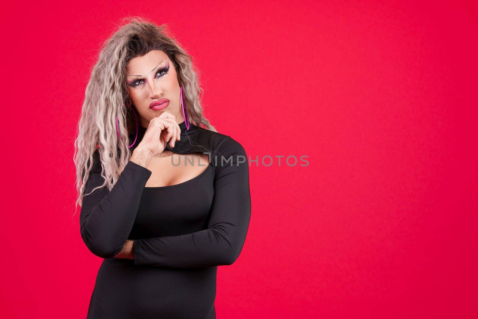 Transgender person with black dress and make up with hand on chin and thoughtful expression in studio with a red background
