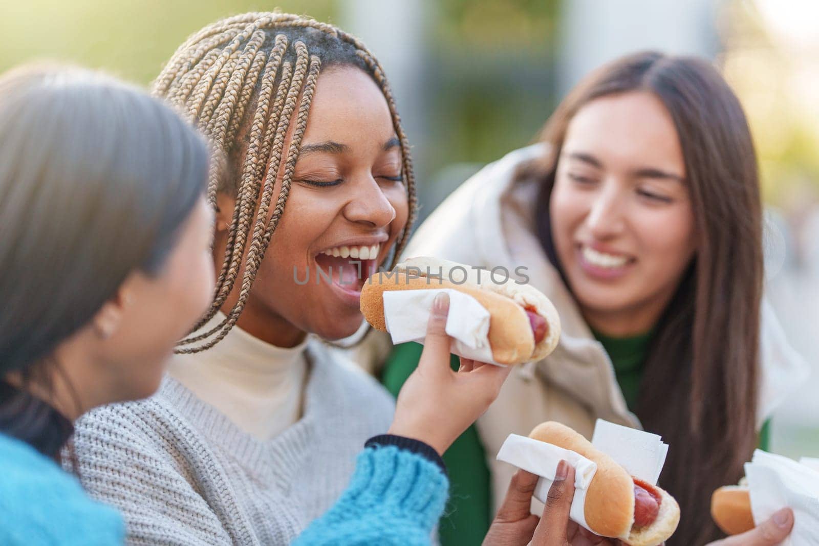 Three multicultural smiling friends eating hot dog in a park