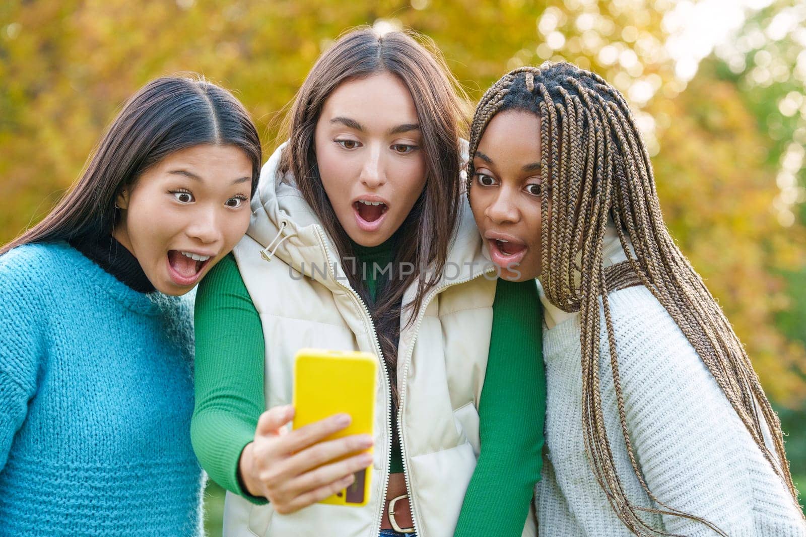 Three surprised multicultural friends using a mobile in a park
