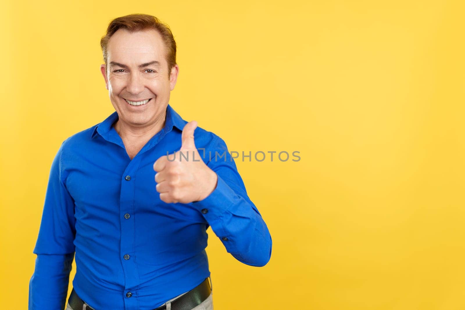 Happy mature man gesturing agreement raising a thumb up in studio with yellow background