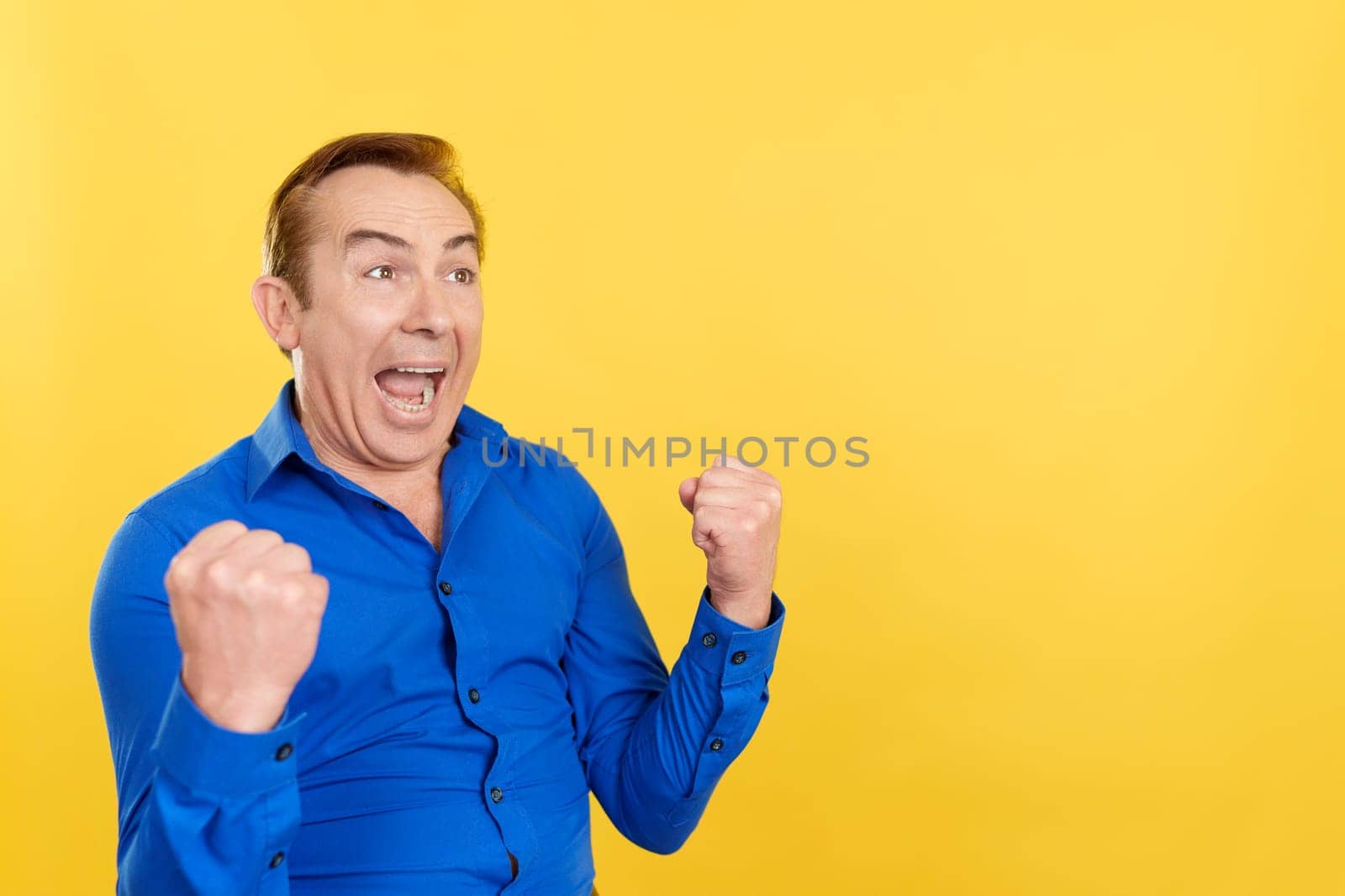 Happy mature man celebrating raising the fists in studio with yellow background