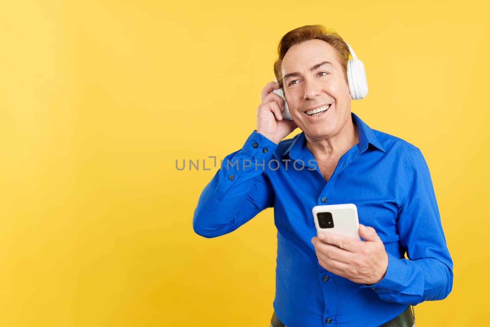 Smiley mature man listening to music with the mobile and headphones in studio with yellow background