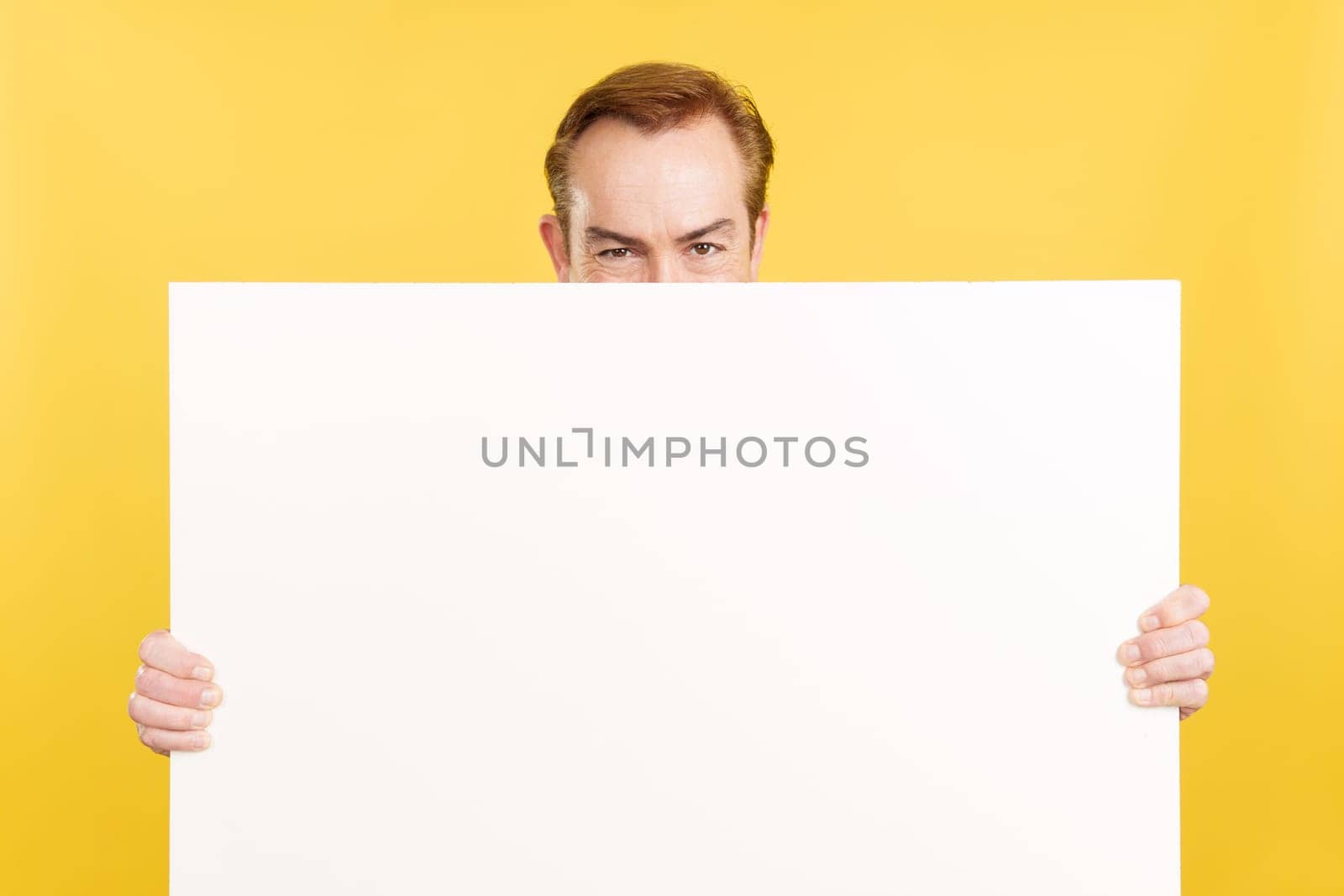 Caucasian mature man hiding behind a blank panel in studio with yellow background