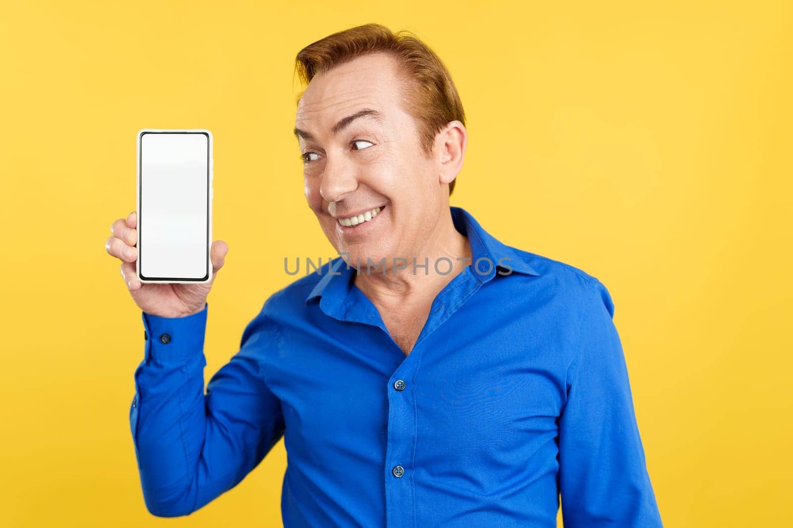 Smiley mature man holding a mobile with a blank screen in studio with yellow background