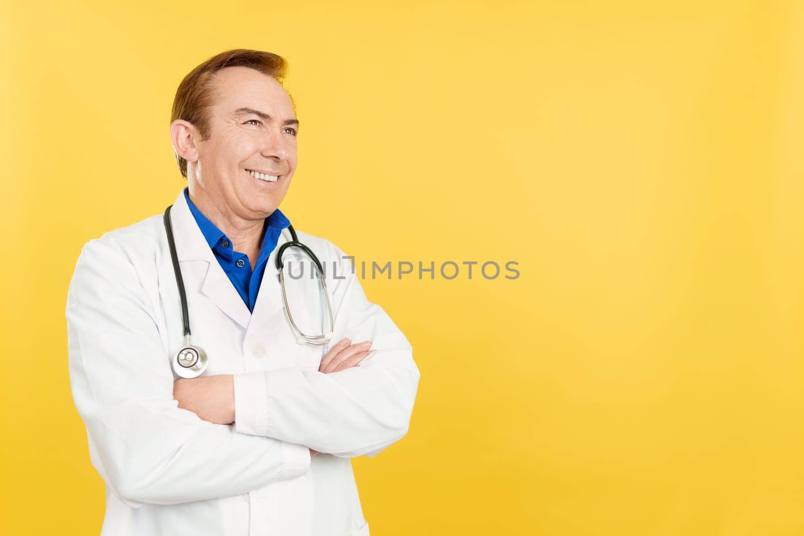 Happy mature caucasian doctor with uniform looking aside distracted with arms crossedin studio with yellow background