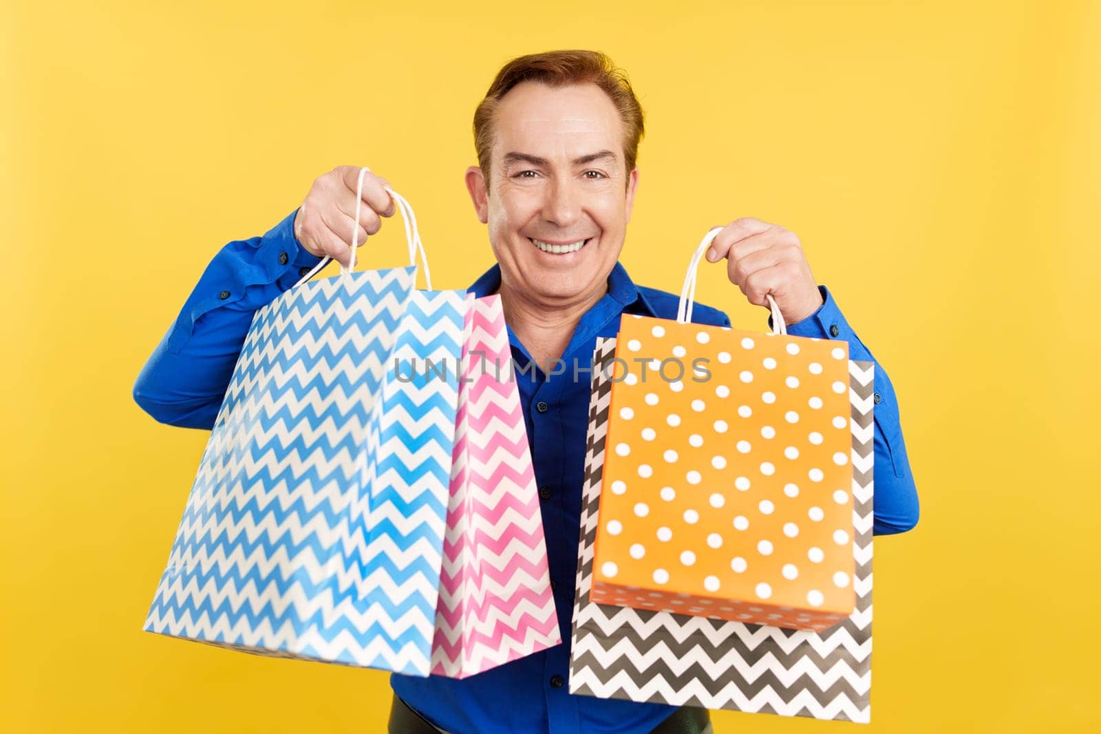 Smiley mature man looking at the camera showing shopping bags in studio with yellow background