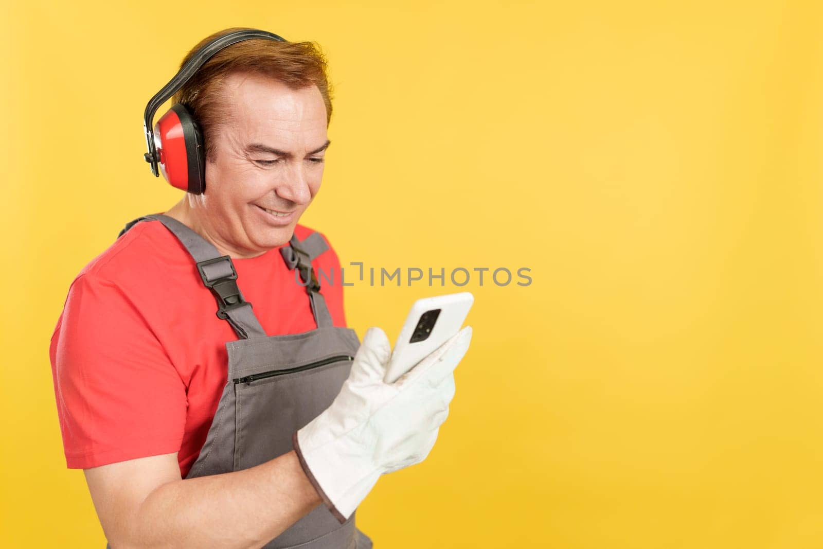 Mature worker in uniform and protective gear using a mobile in studio with yellow background