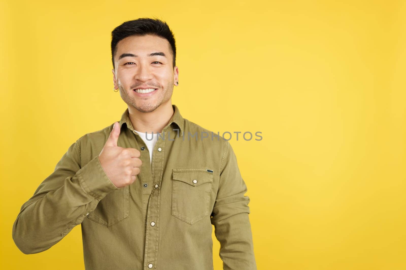 Happy chinese man gesturing agreement raising a thumb up in studio with yellow background